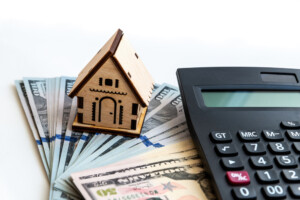 Wooden model of house on stack of cash next to calculator